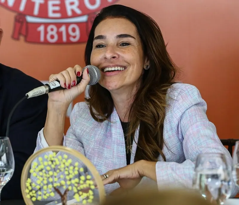 Juliana Benício fala ao microfone em mesa de debate, diante do painel “Niterói 1819”